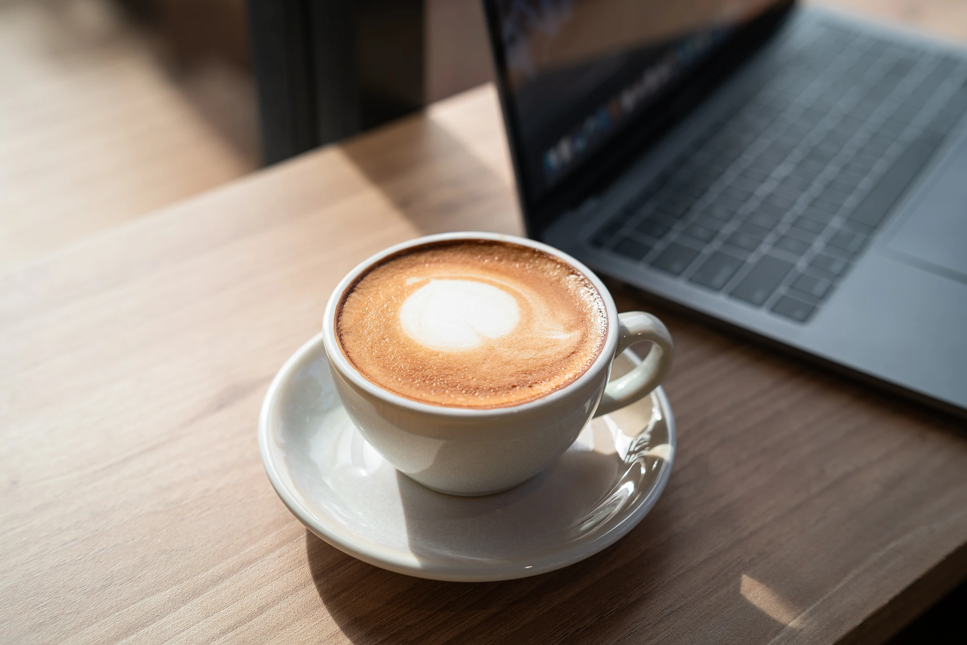 En cappuccino med latte art på ett träbord bredvid en laptop.